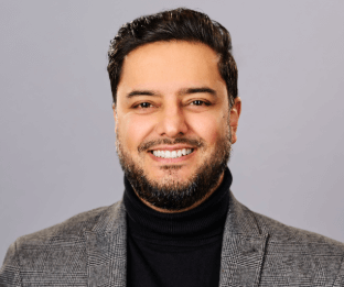 A man with brown hair wearing a dark grey suit jacket and black jumper