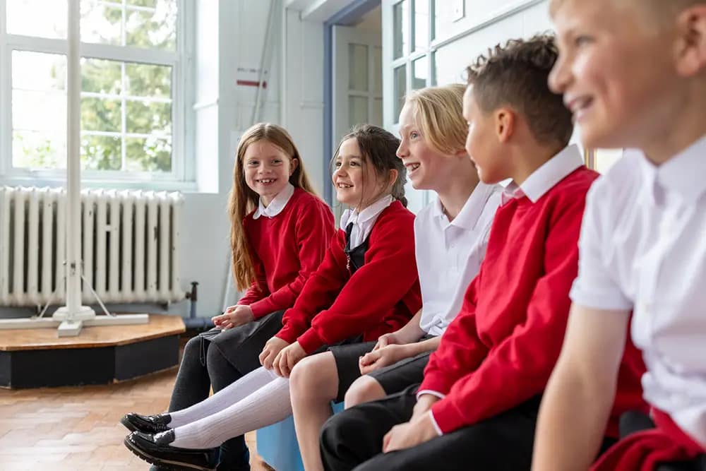 Primary school children sitting together on a bench indoors, smiling and chatting while wearing school uniforms.