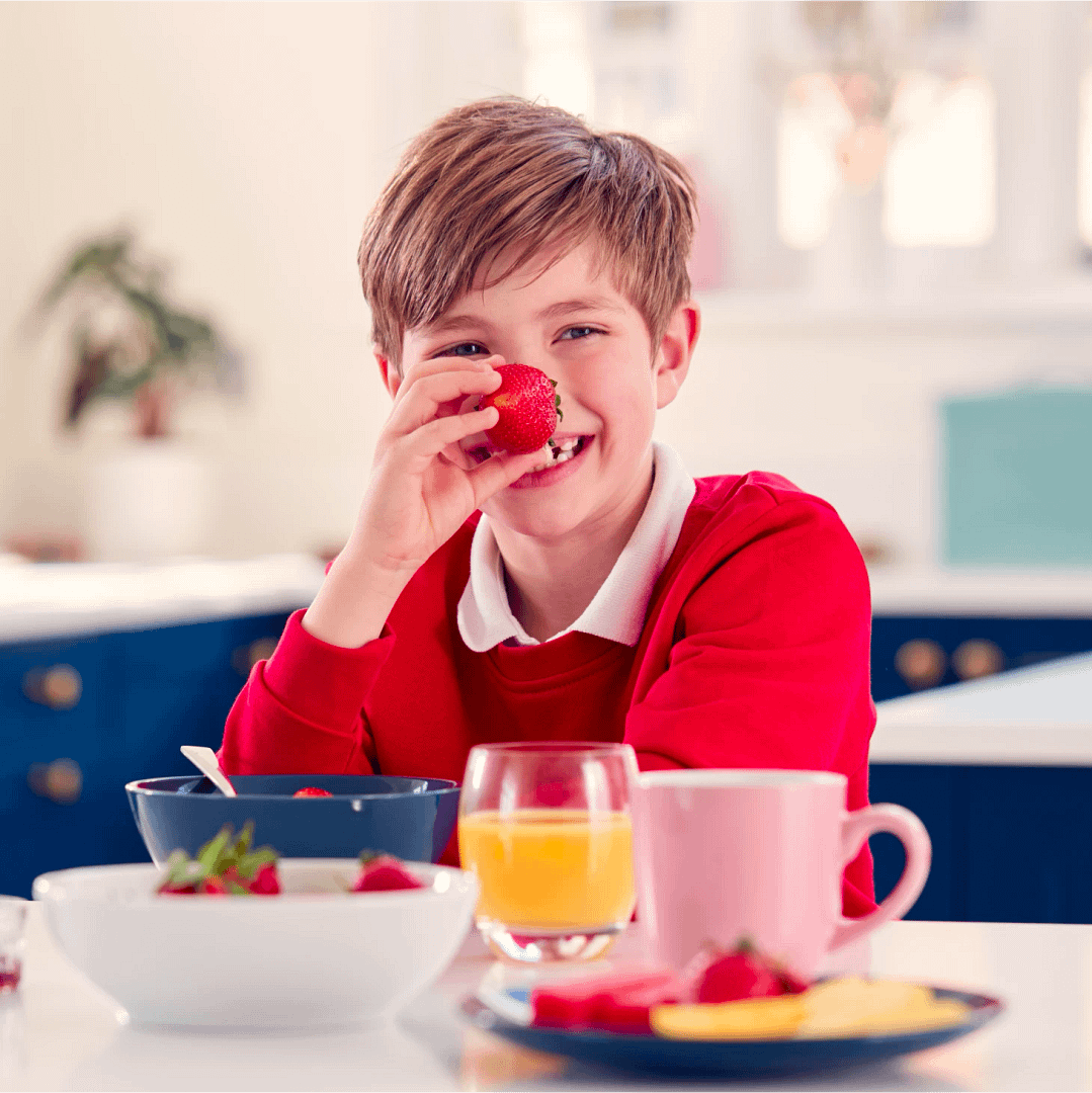 A young boy in a school uniform sitting at a table, eating breakfast with fruit and juice before school.