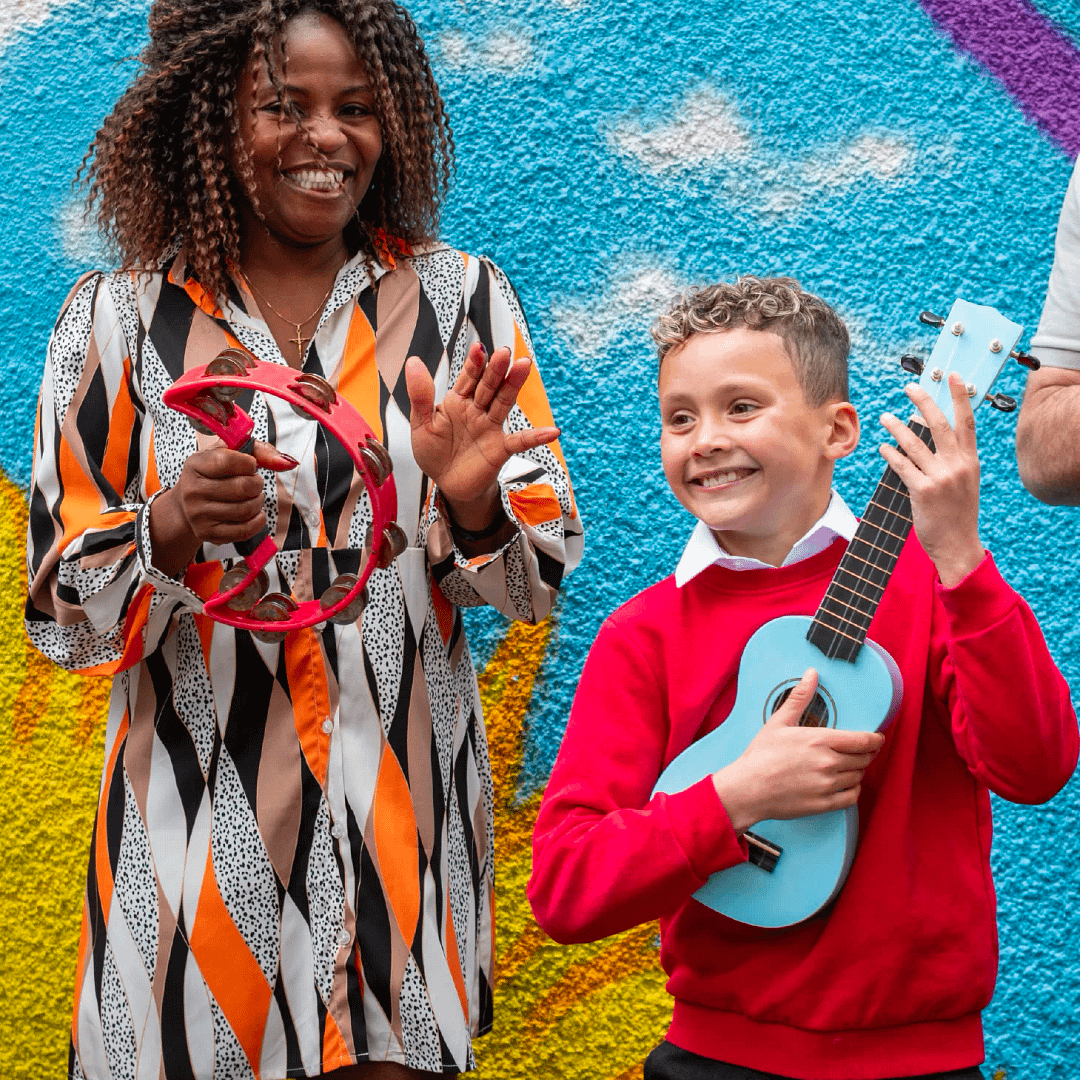 A young primary school boy playing a ukulele next to his teacher, who is playing a tambourine