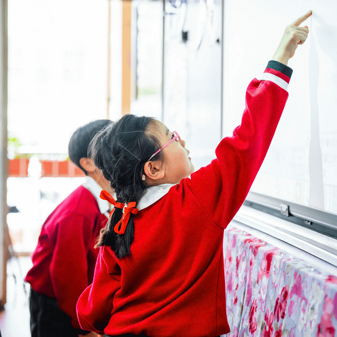Child in a red jumper with glasses and braided hair pointing up at a classroom whiteboard while another student looks on.