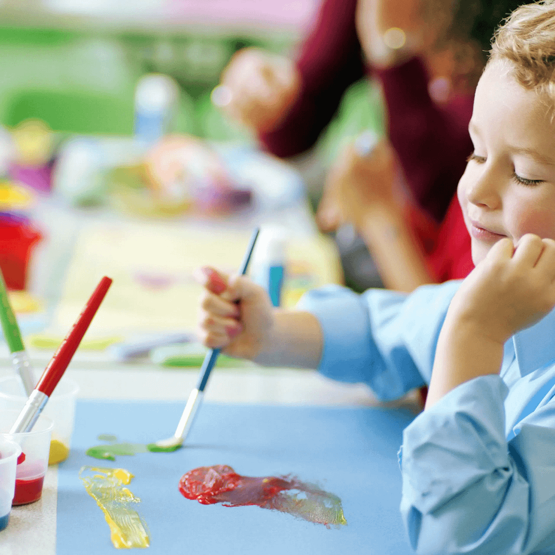 Child in blue shirt paints with red, yellow and green colours on paper during a classroom art lesson.