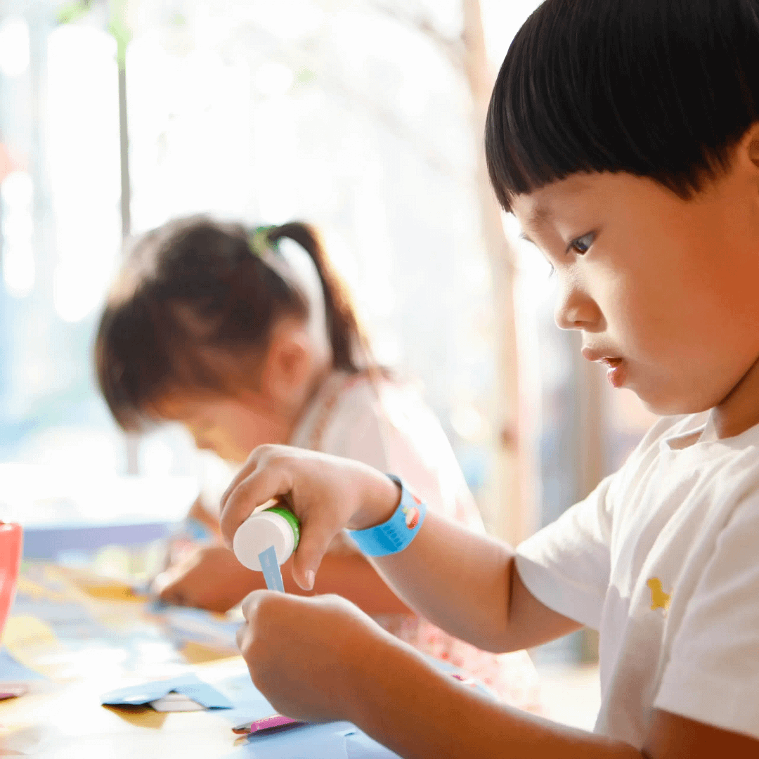 A child carefully applies glue to a piece of paper while another child in the background also works on a craft project at a table.