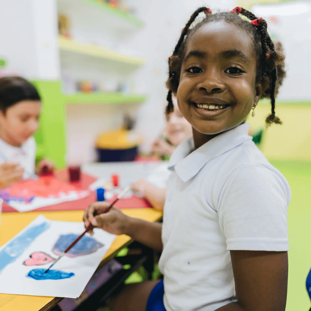 Smiling child in a white shirt paints with blue on paper at a classroom table, with other children painting in the background.