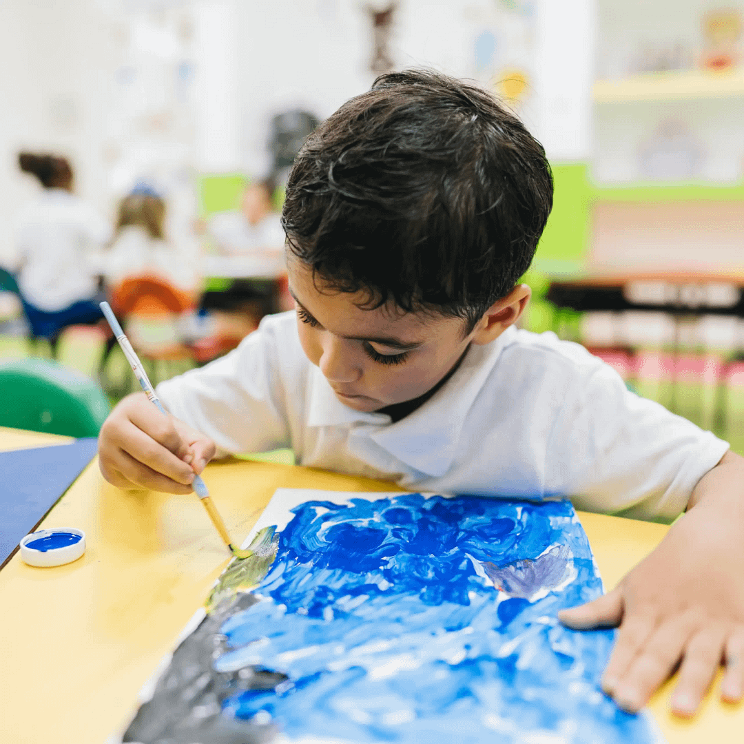 Child concentrating while painting with blue and green colours on paper during an art activity.