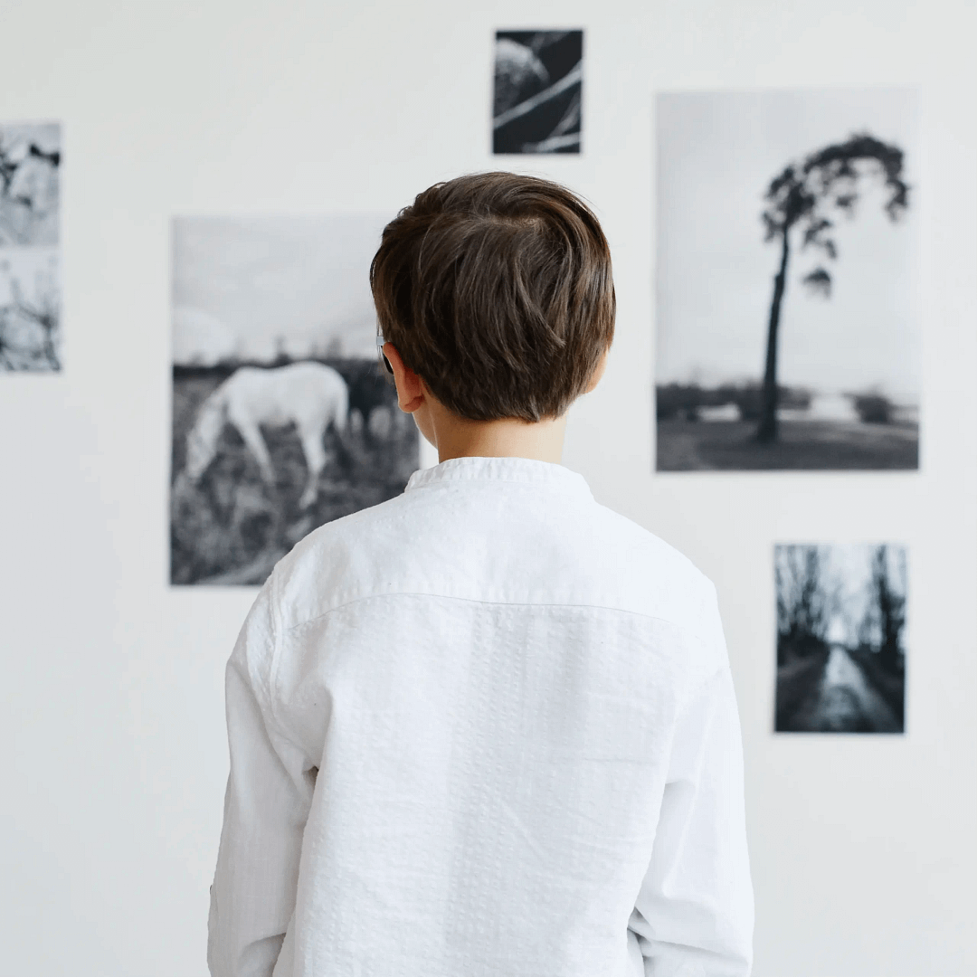 Child wearing a white shirt, seen from behind, observing several black-and-white photographs on a gallery wall, including images of a horse and a tree.