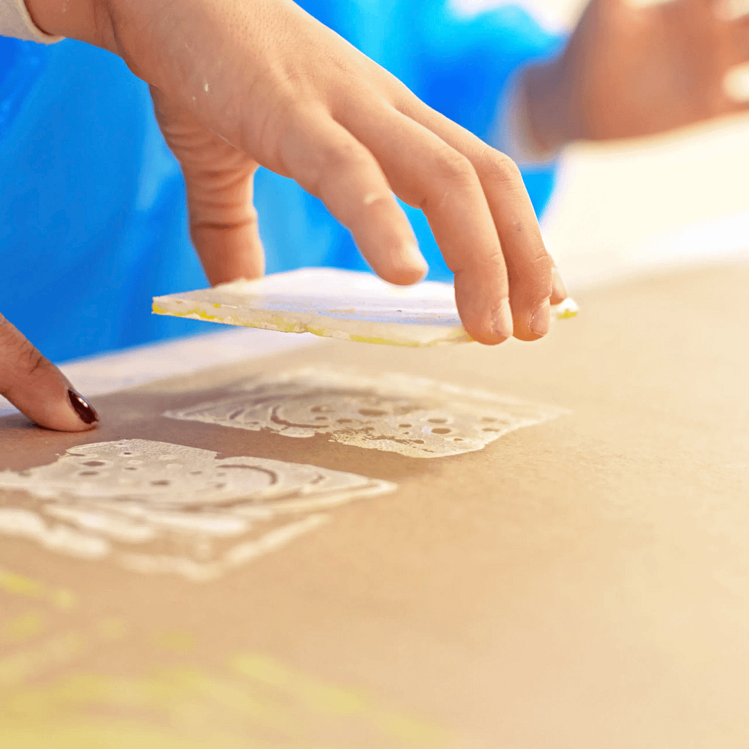 Child’s hand creating block prints on paper, lifting a square stamp to reveal painted decorative patterns in white and yellow.