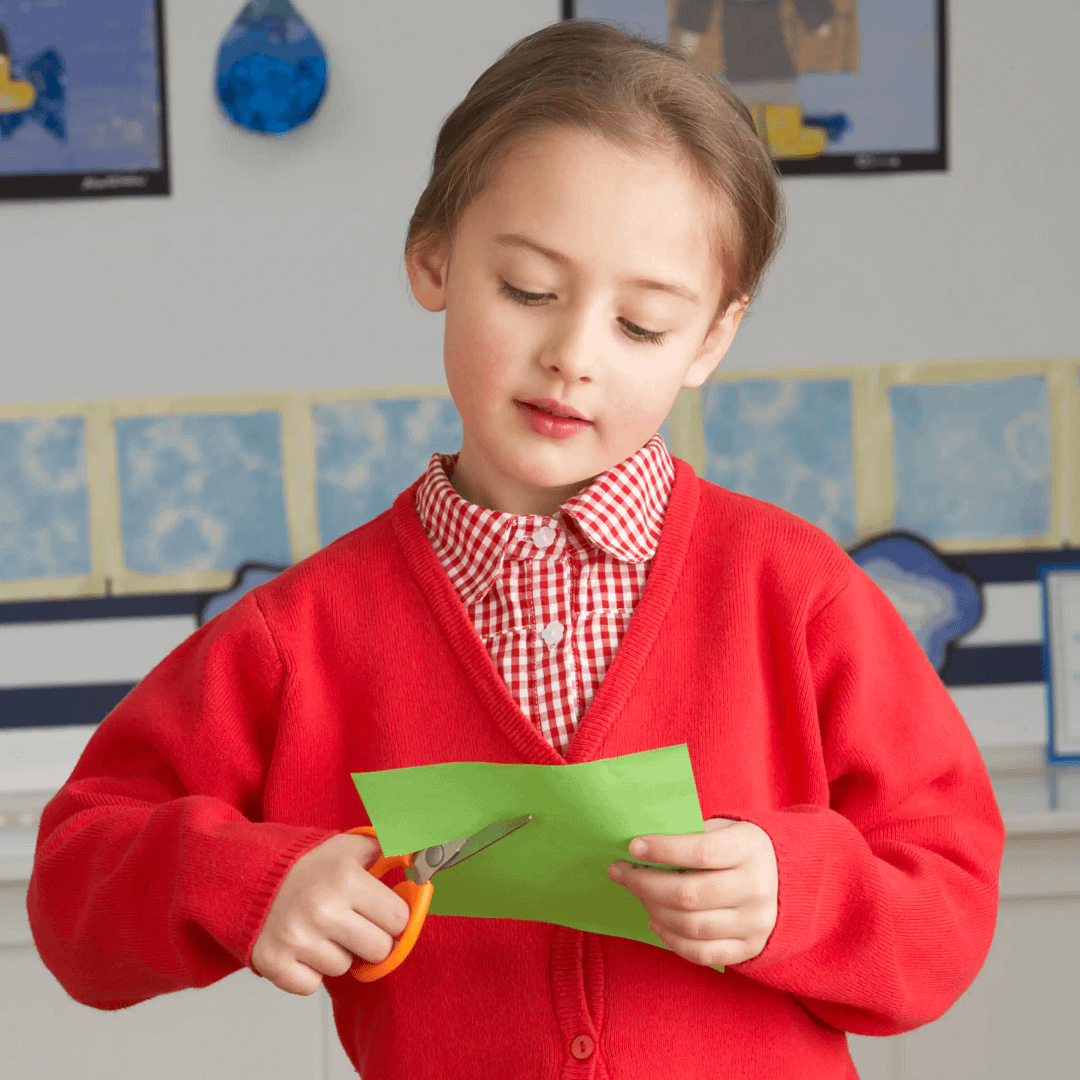 Child in red school uniform cuts green paper with scissors during a classroom craft activity.