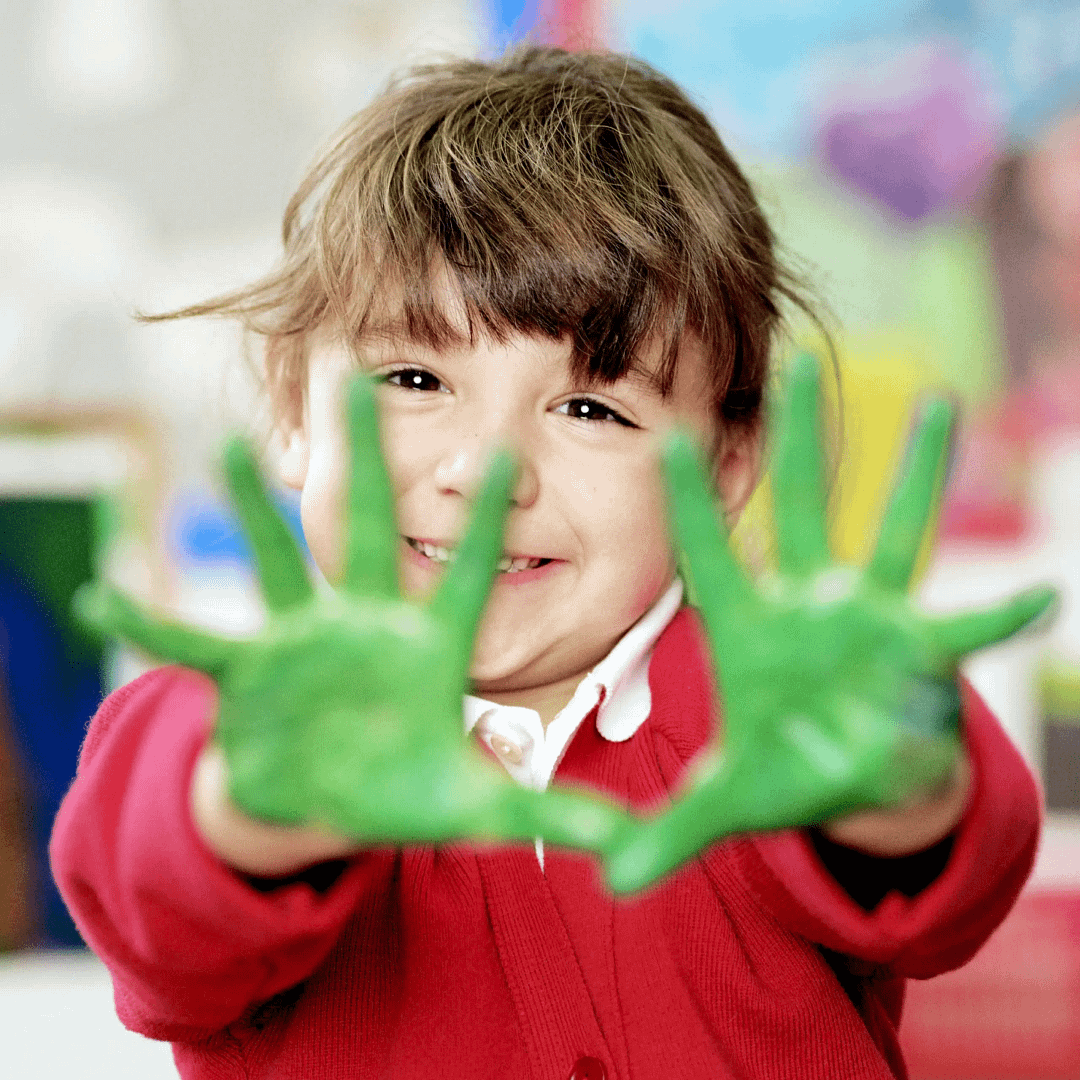 Close-up of a child in a red school jumper smiling and holding out both hands covered in bright green paint.