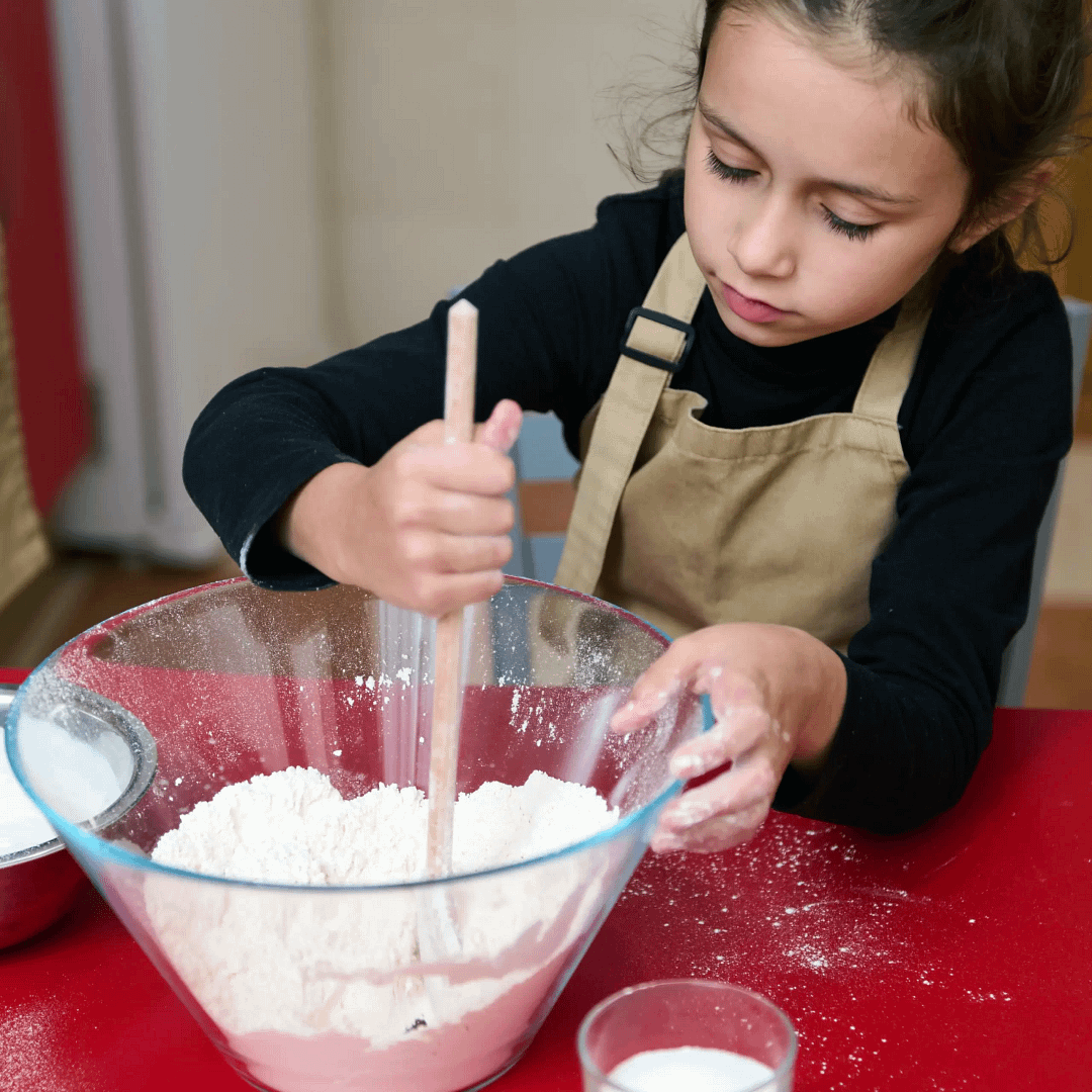 Child wearing an apron mixing flour in a large glass bowl with a wooden spoon.