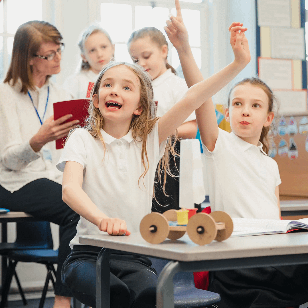 Children in class raising hands with excitement, a small model car on the desk, and a teacher observing in the background.