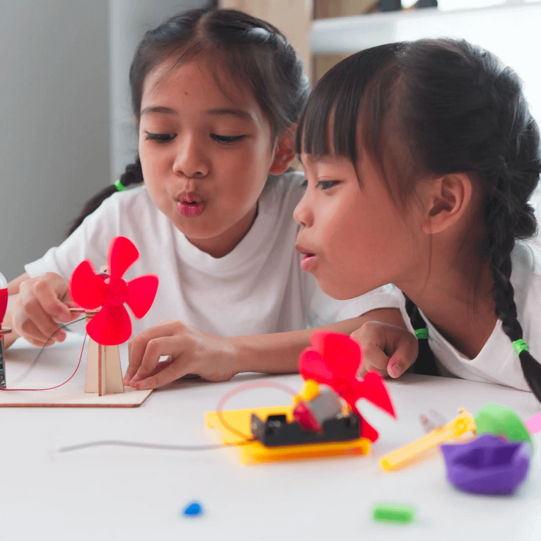 Two children test small red-bladed fan projects on a table, one powered by wires and a motor.