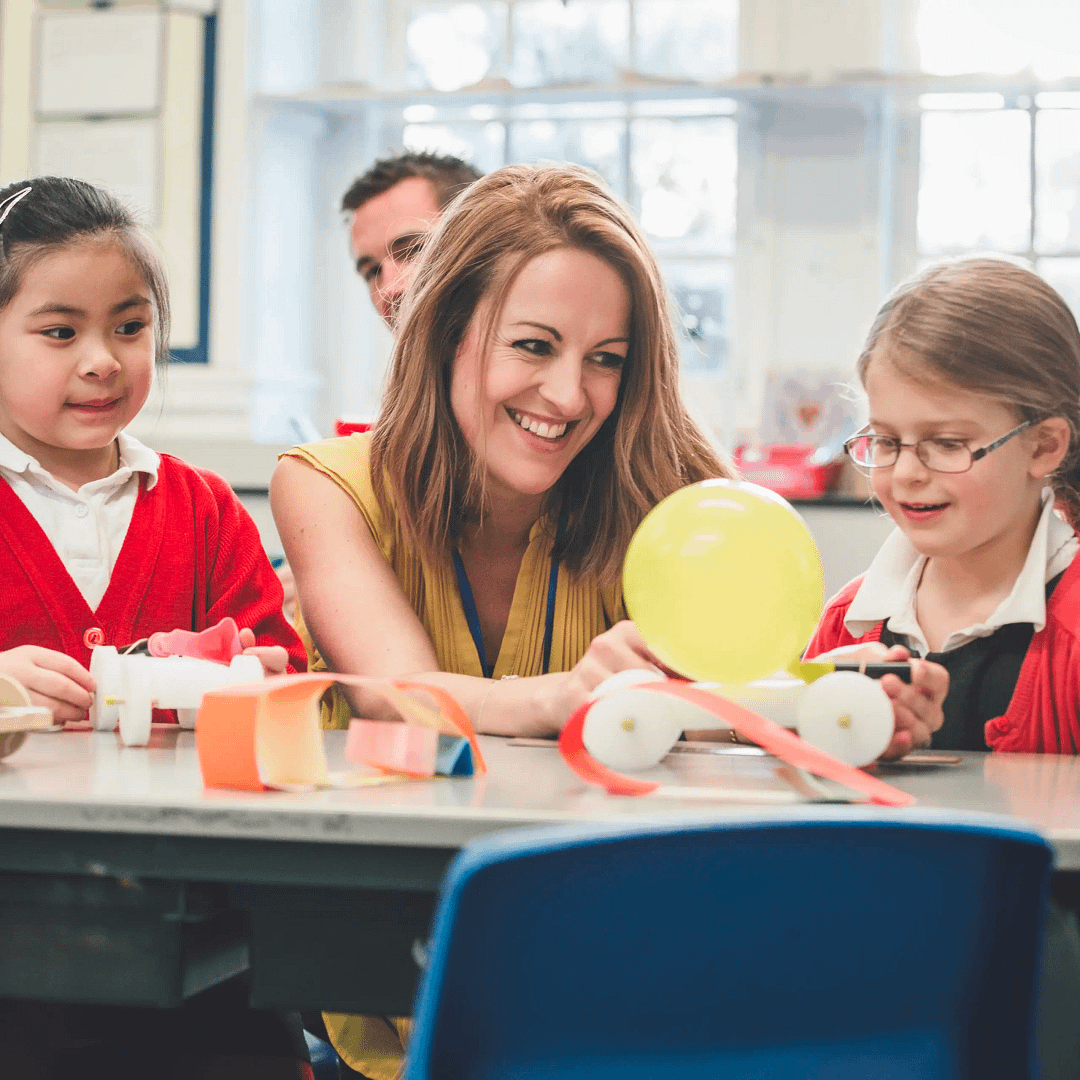 Teacher and two pupils smiling while working on a balloon-powered car project in class.