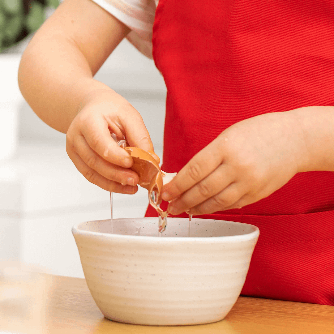 Child in a red apron cracking an egg into a white mixing bowl.