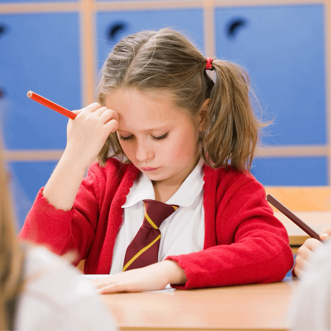 A young girl in a school uniform is sitting at a desk, holding a pencil and concentrating on her work in a classroom setting.