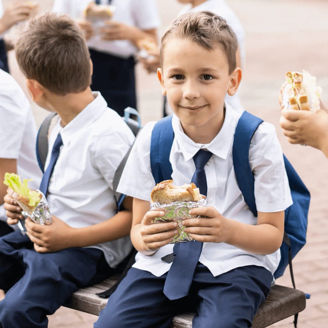 A group of young children wearing school uniform sat outside, eating their lunch together.