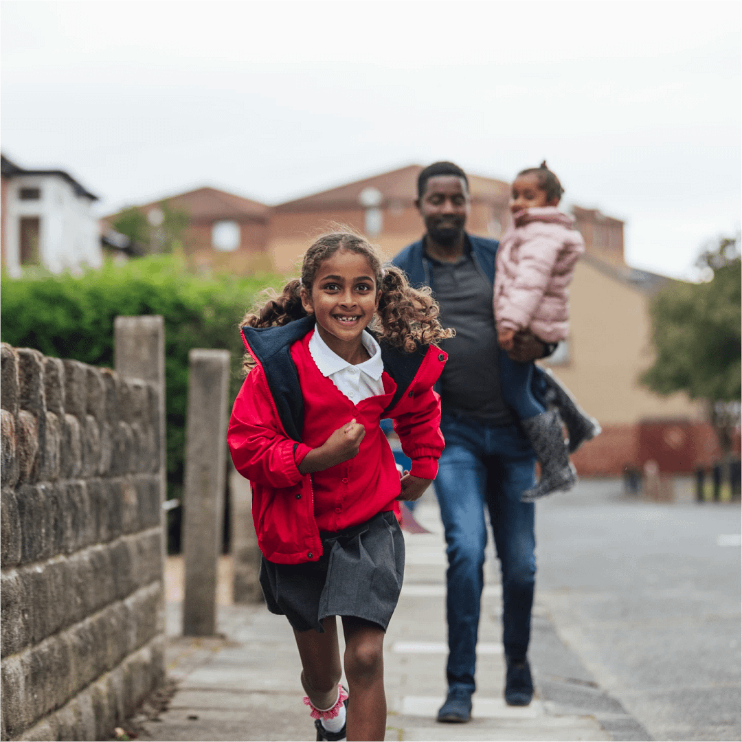 A young girl in a school uniform, running to school with her family following behind her.