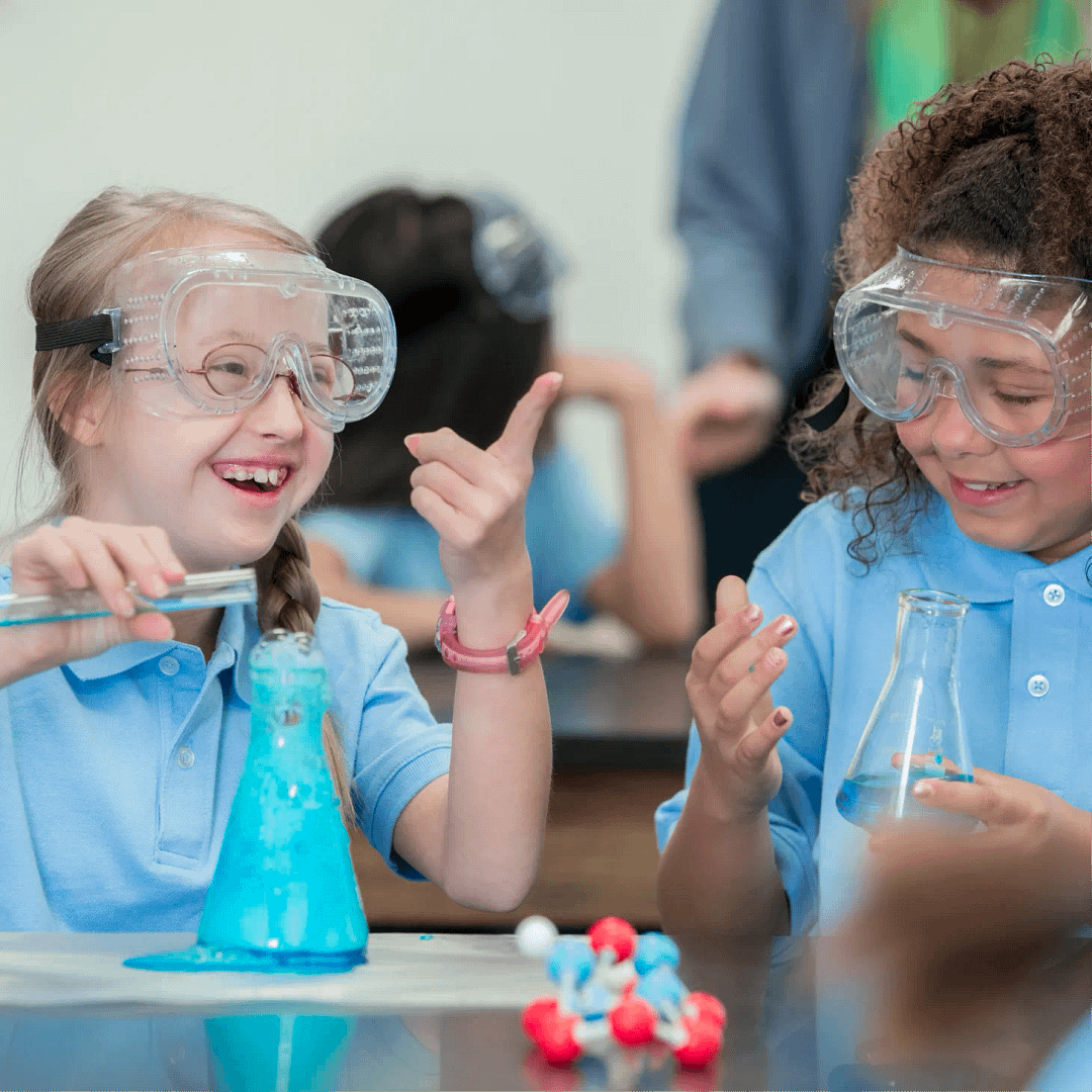 Two young girls in school uniforms wearing goggles are conducting a science experiment in a classroom setting.
