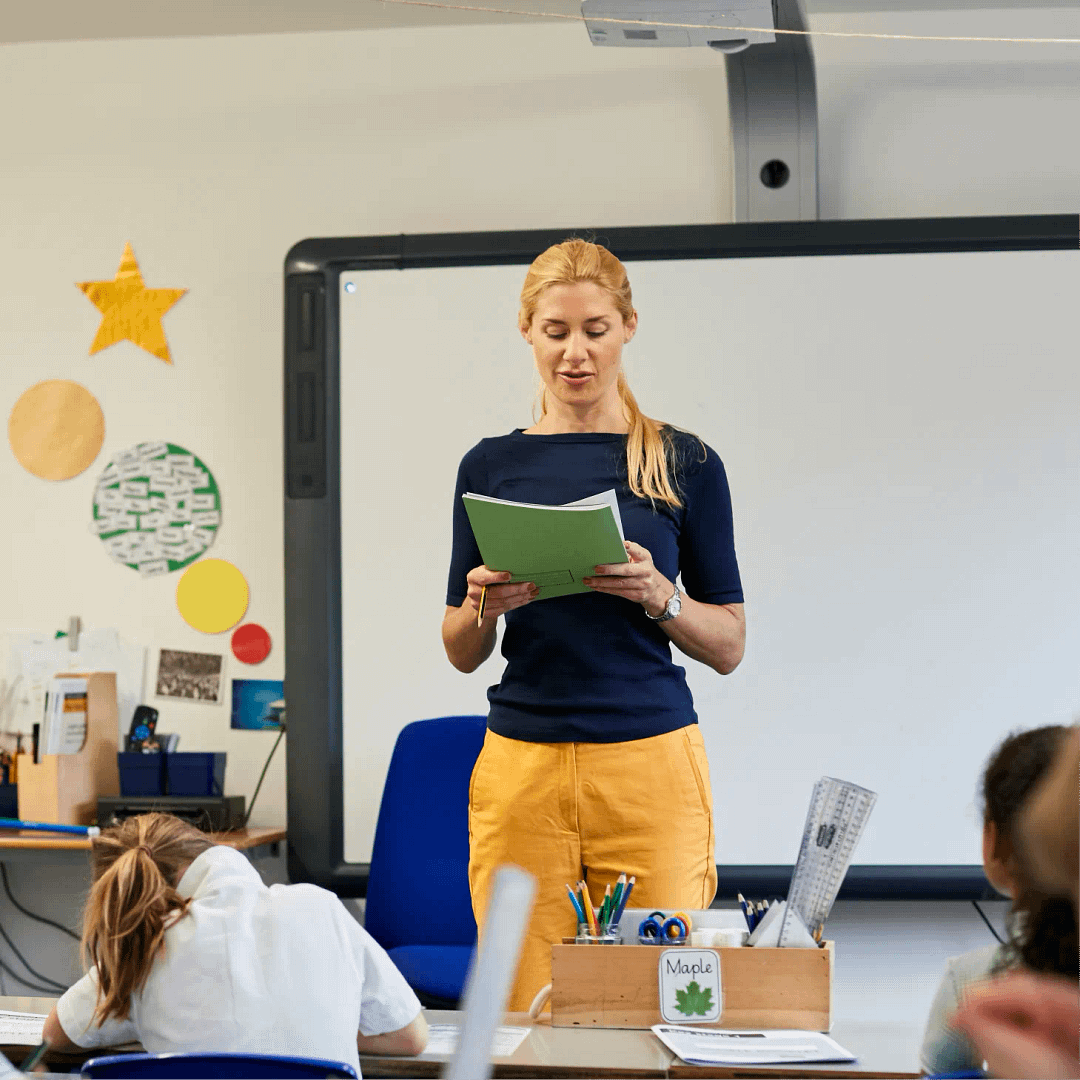 Teacher reading from an extract to the rest of her class, while they are heads-down at their desks writing.