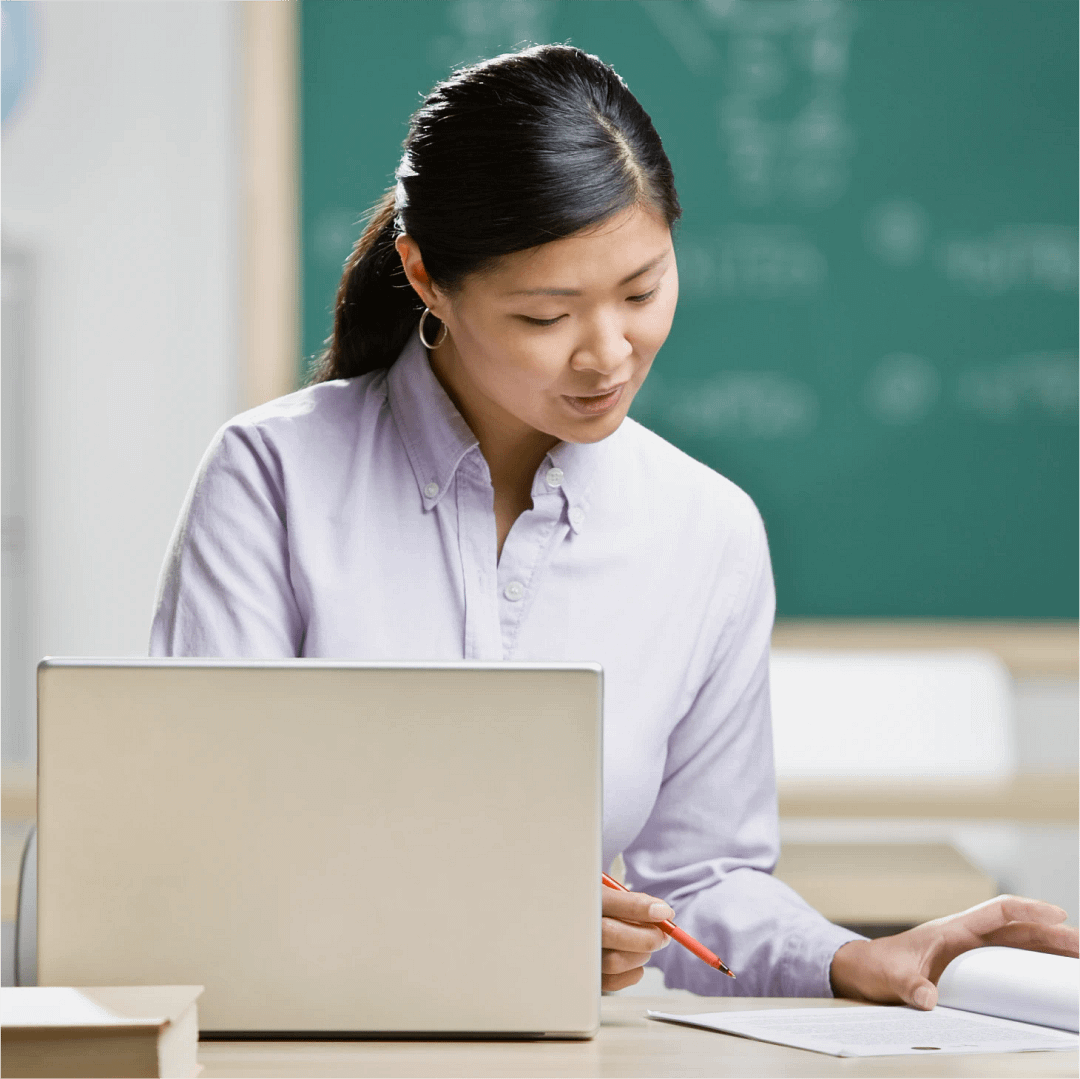A teacher sitting at a desk using a laptop and reviewing paperwork in a classroom, with a chalkboard in the background.
