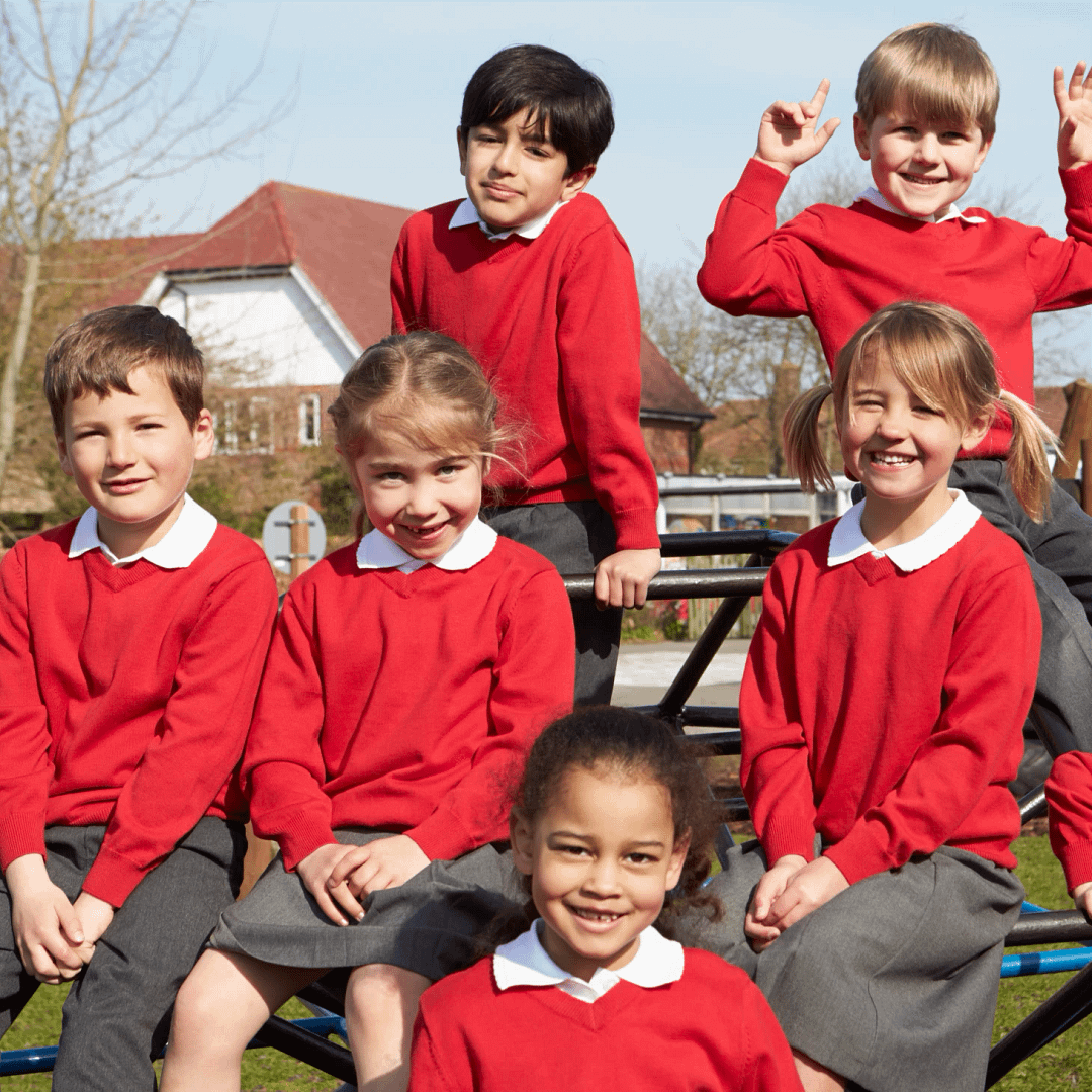 A group of primary school children in red school uniforms smiling and sitting together on playground equipment outside.