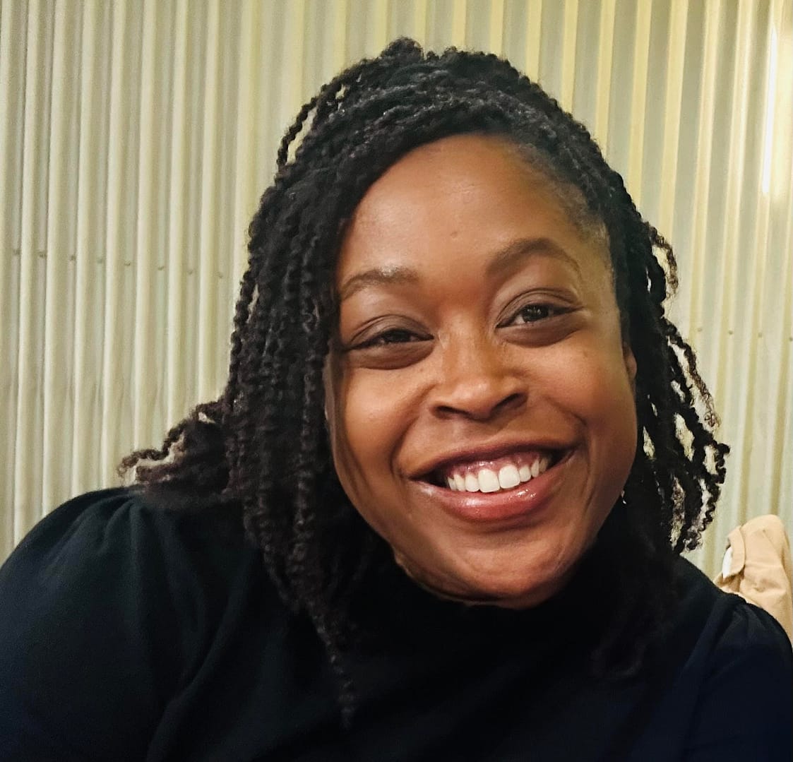 A woman with braided hair smiles warmly at the camera, wearing a dark top, with a light vertical striped background behind her.