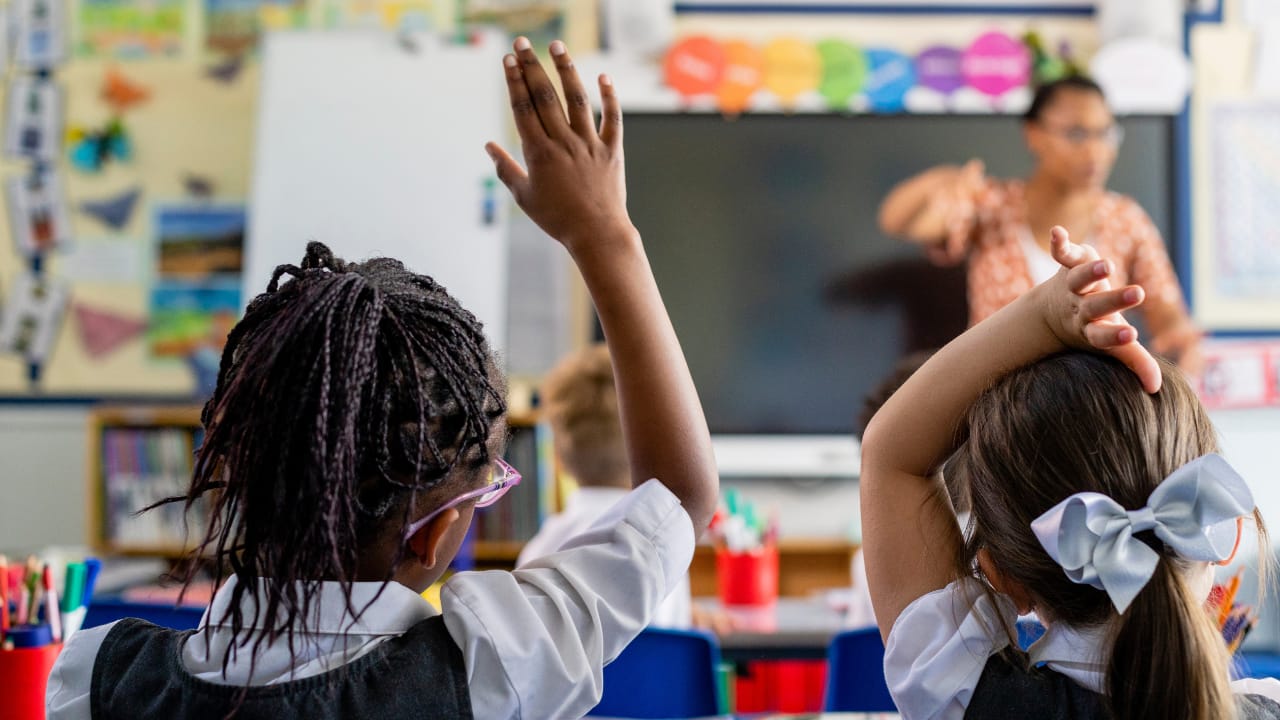 Two primary school pupils sitting at desks with their hands raised during a lesson.
