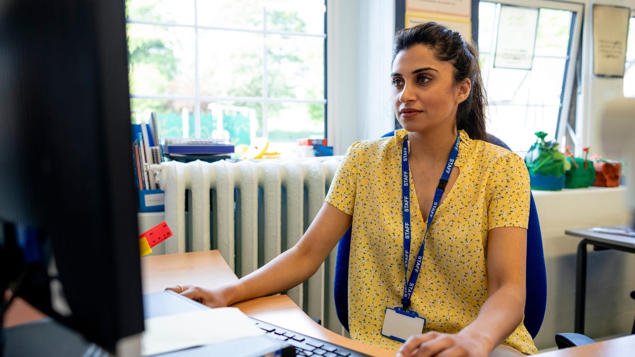 A woman wearing a staff lanyard sits at a desk using a computer in a classroom or office.