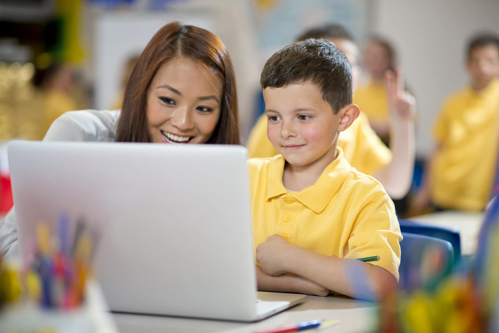 A teacher and a young pupil smiling while looking at a laptop in a classroom.