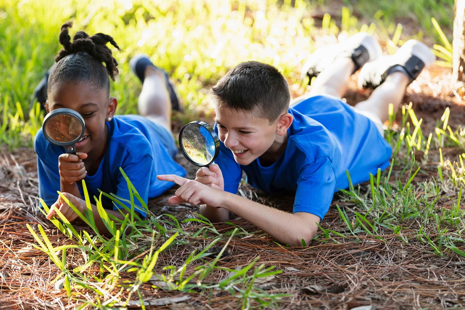 Two children lie on the grass using magnifying glasses to examine plants outdoors.