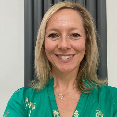 A woman with blonde hair is smiling. She is wearing a green top with gold palm tree patterns and a necklace with a circular pendant. The background features grey vertical blinds.