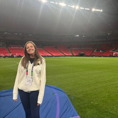 A woman standing on a football field, smiling and wearing a white jacket with a lanyard around her neck, with empty red stadium seats in the background.