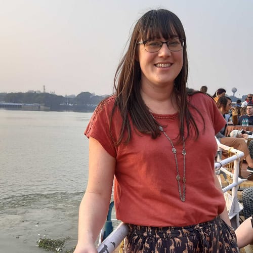 A woman with long dark hair and glasses is smiling while standing on a pier. She is wearing an orange top and patterned trousers, with a necklace. The background shows a body of water and other people on the pier.