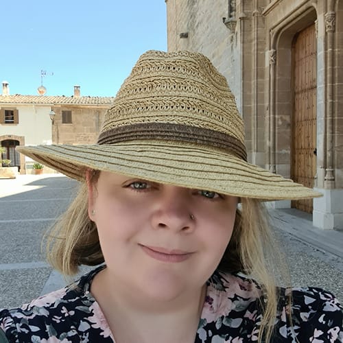 A woman with light hair is smiling and wearing a wide-brimmed straw hat. She is standing in a sunlit courtyard with historic buildings in the background.