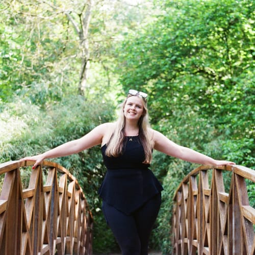 A woman with long, light hair is standing on a wooden bridge in a lush, green park. She is smiling and wearing a black outfit with sunglasses resting on her head, enjoying the natural surroundings.