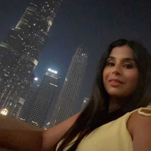 A woman with long, dark hair is smiling and wearing a light yellow top. She is seated outdoors at night with a city skyline featuring tall, illuminated buildings in the background.