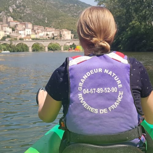 A person with blonde hair is seen from behind, paddling a green kayak on a river. They are wearing a purple life vet. In the background, there is a stone bridge and a small town nestled among green hills.