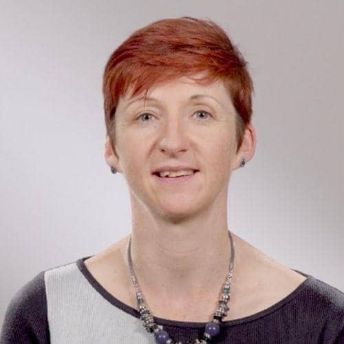 A woman with short, red hair is smiling. She is wearing a grey and black top with a beaded necklace. The background is a neutral grey.