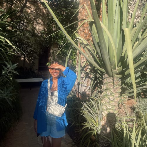 A woman is standing outdoors in a sunny, lush garden with large green plants. She is wearing a blue shirt, white top, and shorts, with a small white bag across her body. She is smiling and shielding her eyes from the sun with her hand.