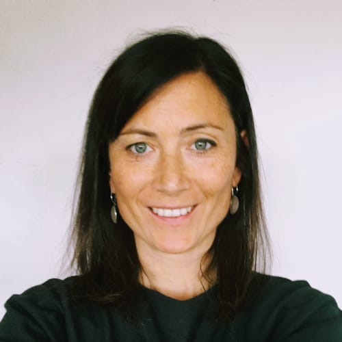 A woman with dark hair is smiling. She is wearing a black top and earrings. The background is a plain light colour.