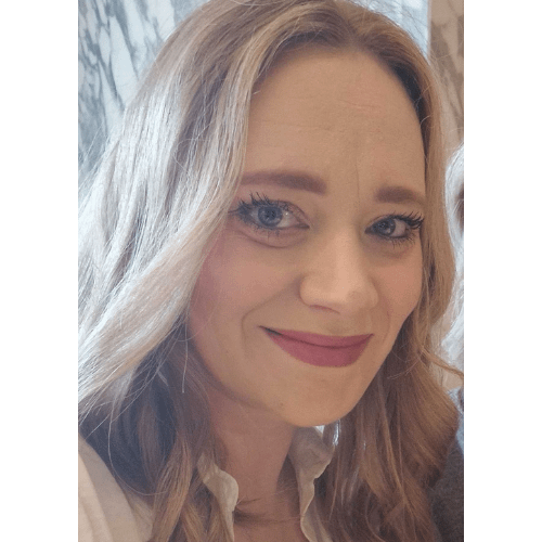 A woman with light brown /blonde hair is smiling. She is wearing a white blouse and a necklace with a pendant. The background features a marbled pattern.