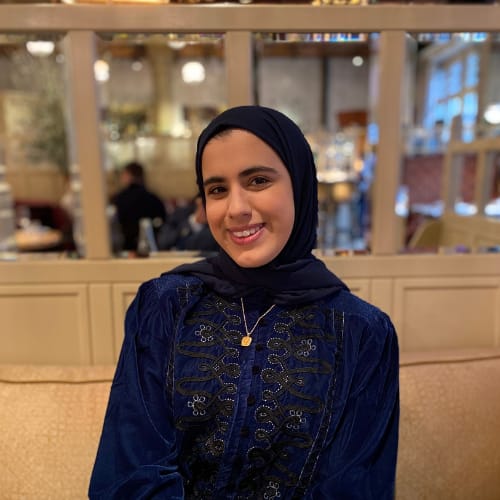 A woman wearing a dark blue hijab and a matching dark blue embroidered top is smiling. She is seated indoors with a blurred background of a warmly lit restaurant.