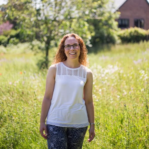 A woman with curly hair and glasses standing in a sunny, lush field, wearing a white sleeveless top and patterned trousers, smiling at the camera.