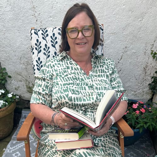 A woman with glasses sitting outdoors in a patterned chair, wearing a green and white dress, holding an open book with additional books on her lap and a potted plant in the background.