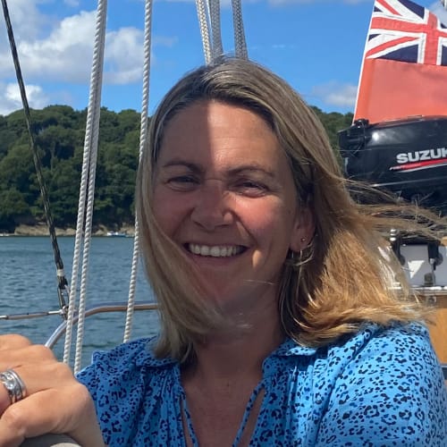 A woman with blonde hair is smiling while sitting on a boat. She is wearing a blue patterned top. The background shows a body of water, green trees, and part of the boat with a British flag.