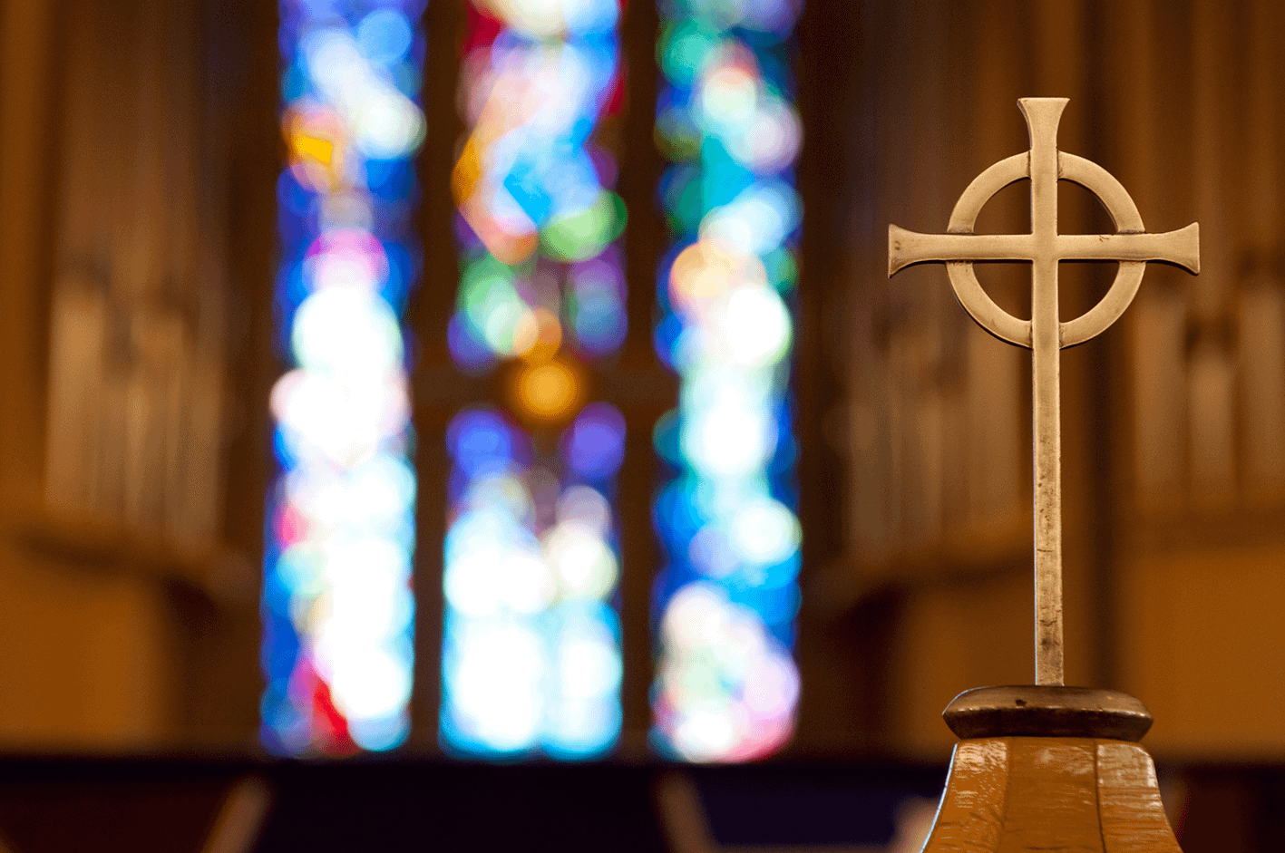 A cross with a blurred stained glass window in the background, creating a peaceful, spiritual atmosphere.