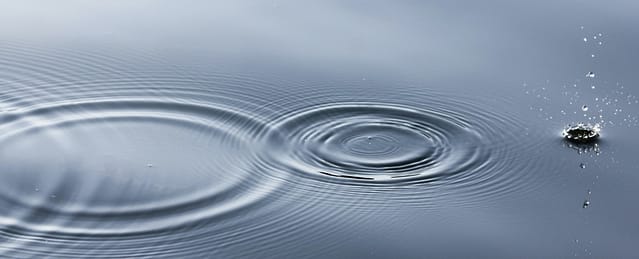 Ripples on a calm water surface, with a single droplet creating concentric circles and splashing nearby.