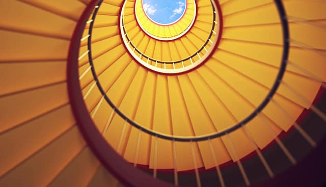 Upward view of a spiral staircase with yellow steps and red railing, leading to a circular opening with a view of the sky.