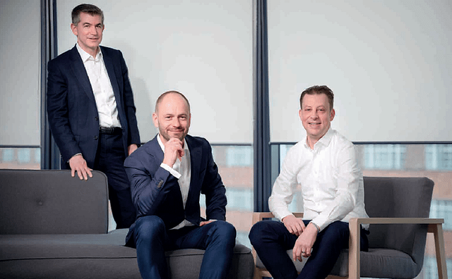 Three men in business attire pose thoughtfully in a modern office space.